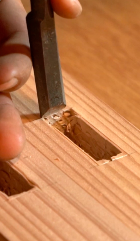 Close-up of a hand carving a rectangular mortise in a piece of wood using a chisel.