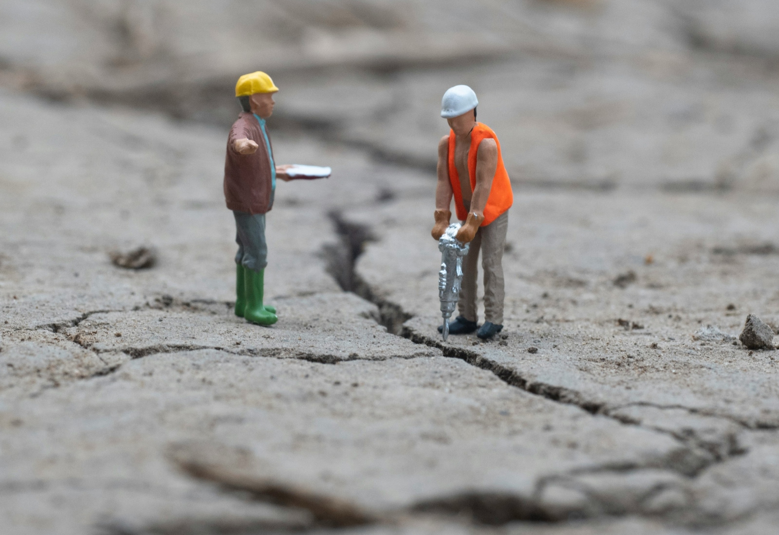 Miniature figures of two construction workers on cracked dry ground, one operating a jackhammer and the other holding a clipboard.