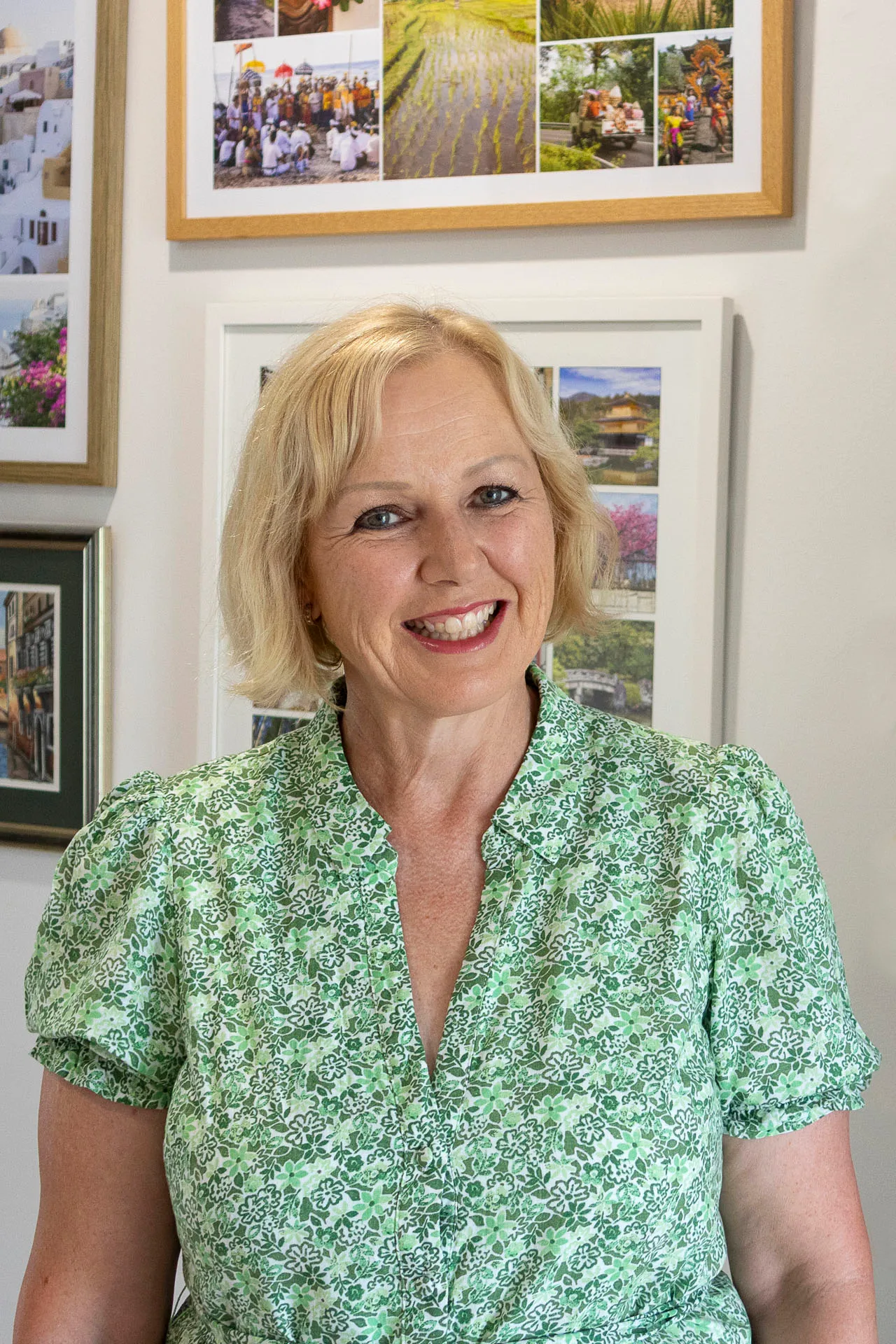 Smiling middle-aged woman with short blonde hair wearing a green floral blouse standing in front of a wall with framed travel photos.