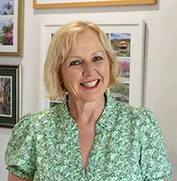 Smiling woman with short blonde hair wearing a green patterned blouse standing in front of framed colorful artwork.