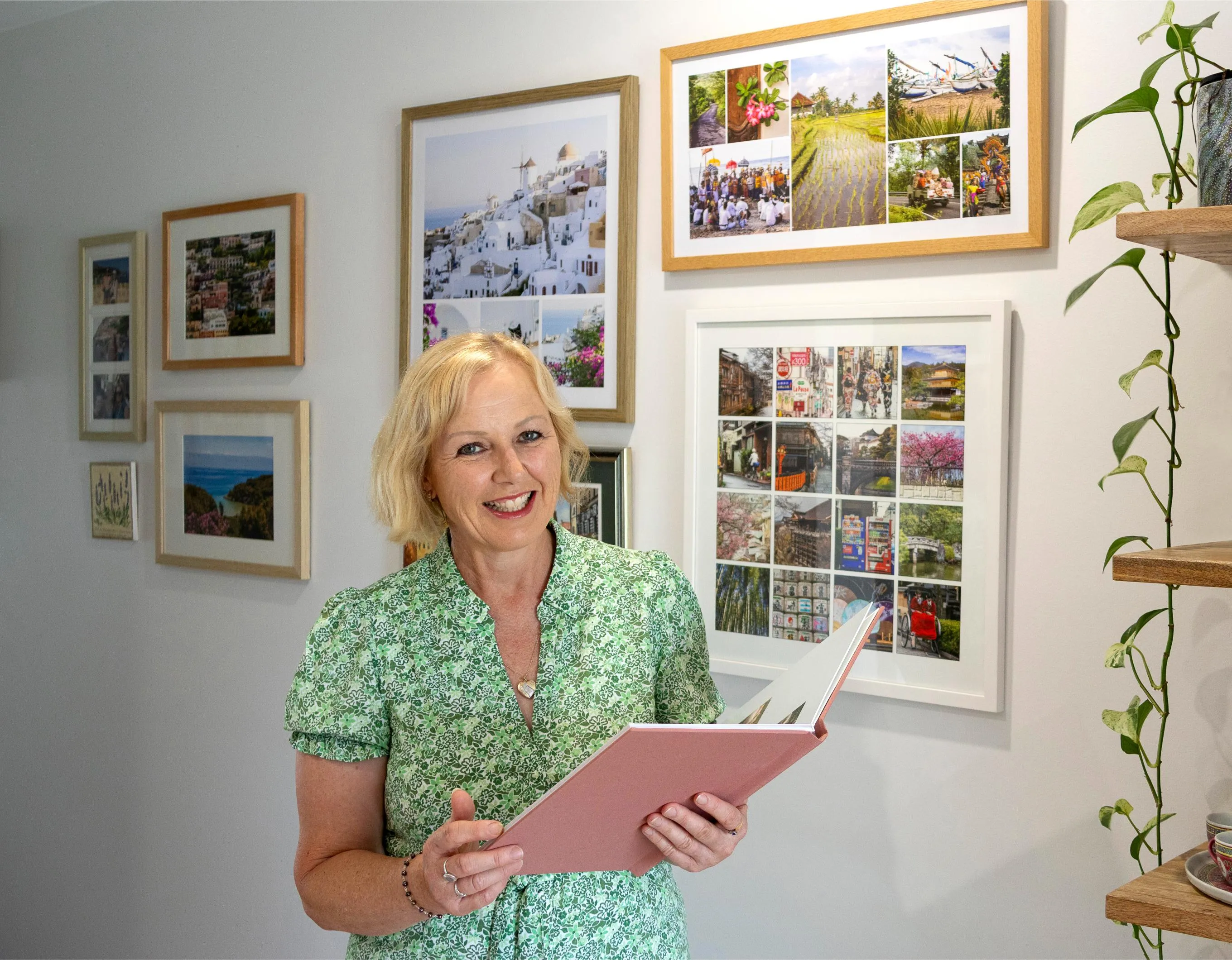 Smiling woman in a green floral dress holding an open pink photo album in a room with framed collage pictures on the wall.