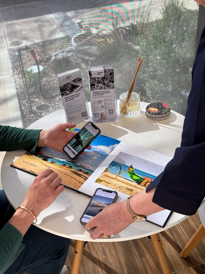 Two people at a round white table viewing a photo book and comparing images on their smartphones in natural light.