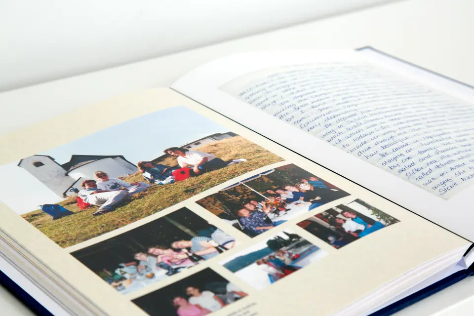 Open photo album on a table showing a group of people sitting on grass near white buildings and various other group photos, with a handwritten note on the opposite page.
