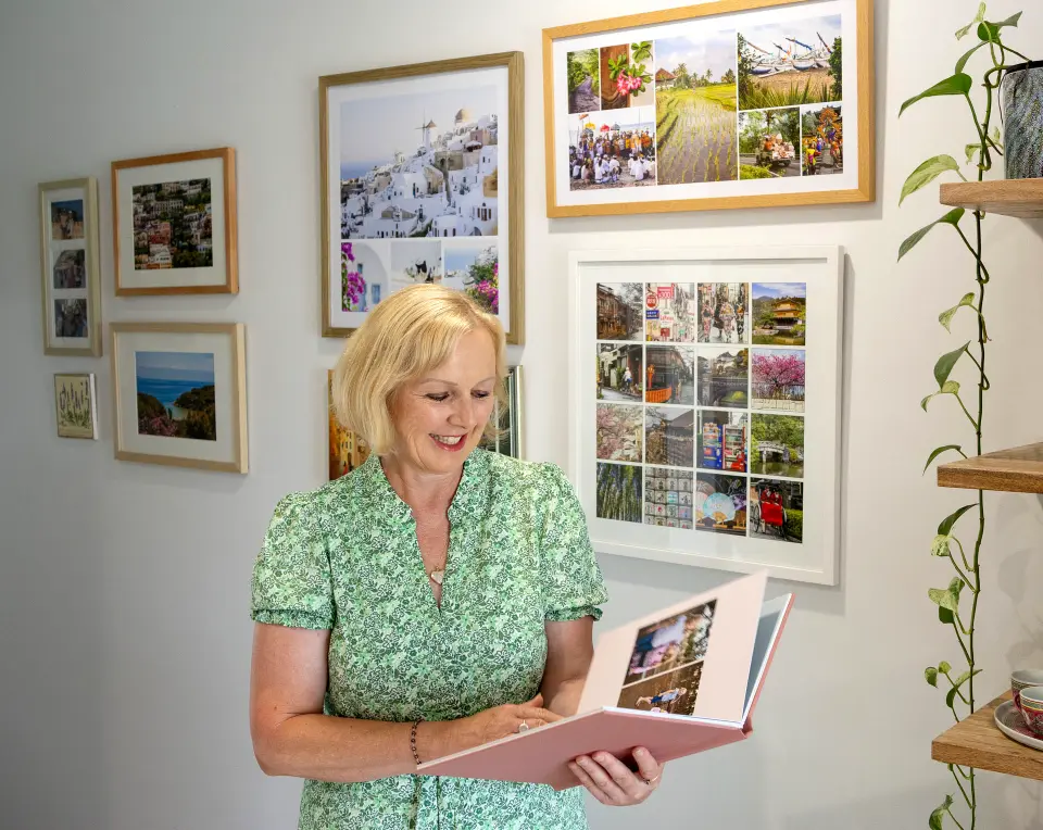 Smiling blonde woman in a green floral dress looking at a photo album in a room decorated with multiple framed travel photos and a hanging plant.