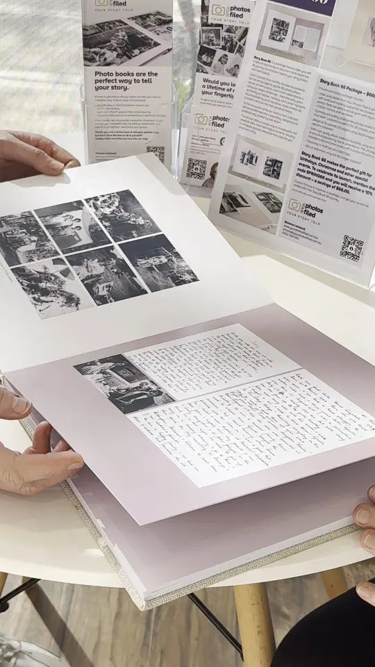 Hands holding open a photo book with black and white photos and handwritten text, displayed on a round white table with promotional flyers for photo books in the background.