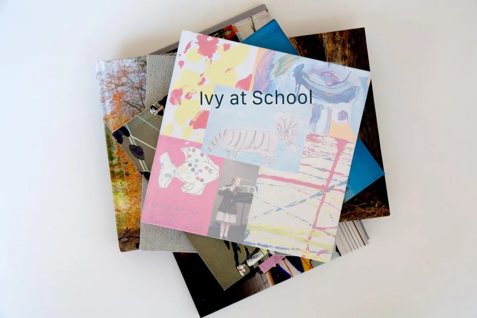 Stack of photobooks with the top one titled 'Ivy at School' featuring colorful child-like drawings and a small photo of a girl playing a flute.