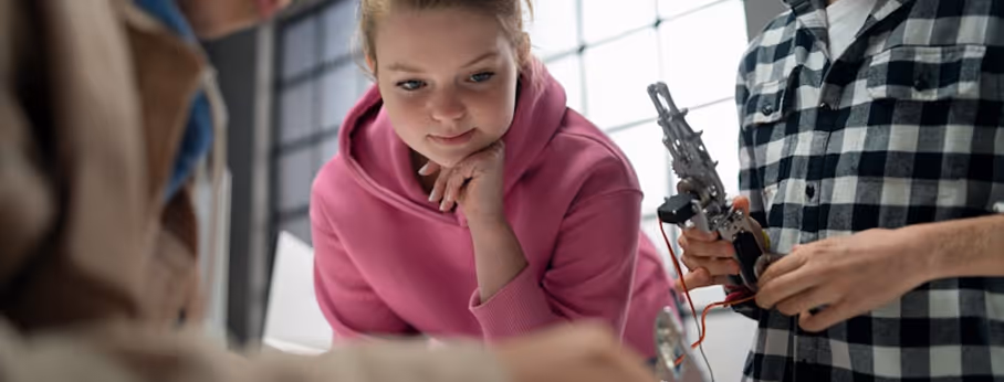 Young woman in pink hoodie observing a robotic hand held by a person in a checkered shirt during a robotics project.
