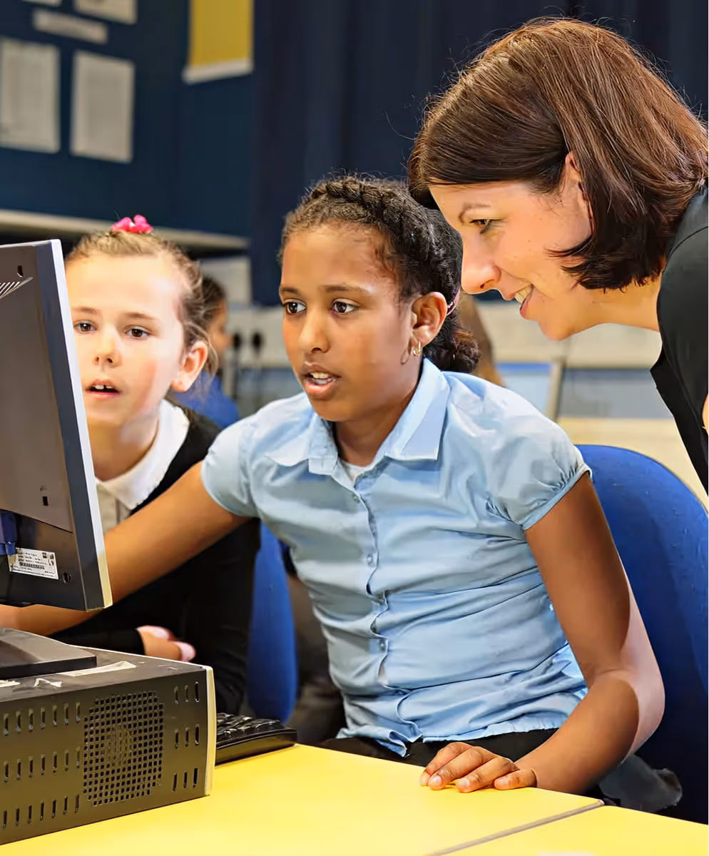 Teacher assisting two young students working on a desktop computer in a classroom.
