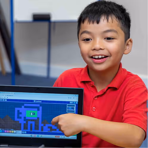 Smiling boy in a red shirt pointing at a pixel art character displayed on a laptop screen.