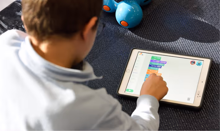 Young boy interacting with a tablet displaying block-based coding software, with a blue robot toy nearby.