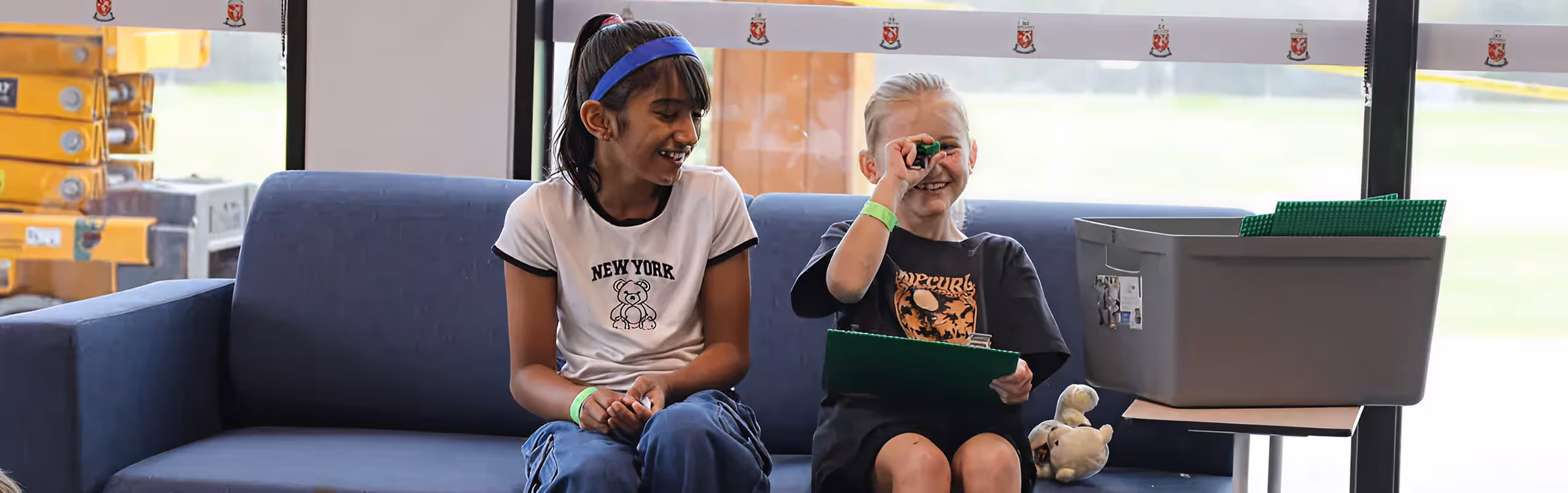 Two children sitting on a blue couch, smiling and playing with a small green object and holding a clipboard.