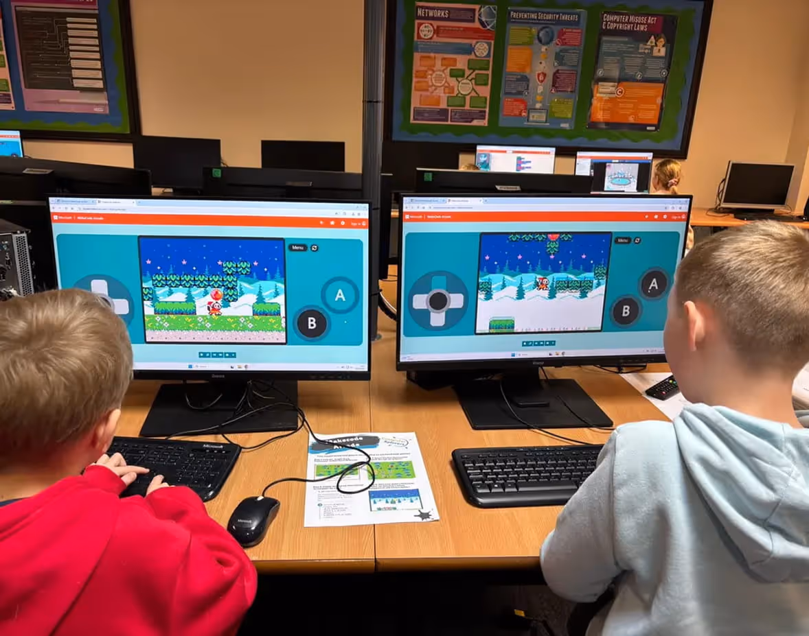 Two children seated at desks using computers with a retro-style platform video game displayed on both monitors.