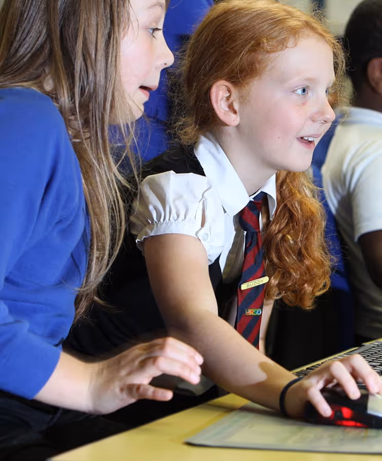 Two girls working together at a computer, one with red hair in a school uniform and the other with brown hair in a blue top.
