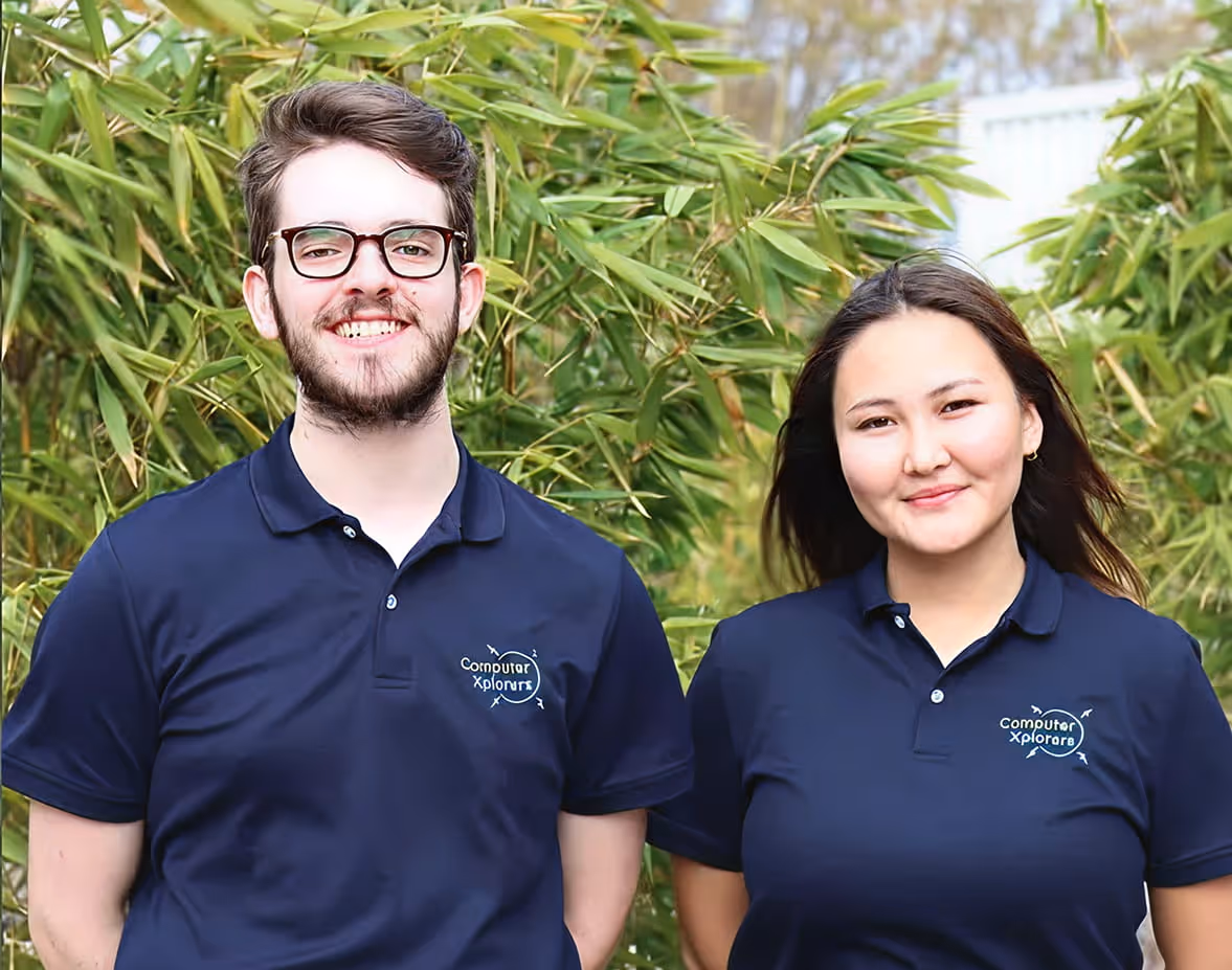 Smiling young man and woman wearing navy blue polo shirts with Computer Xplorers logo, standing outdoors with green foliage background.