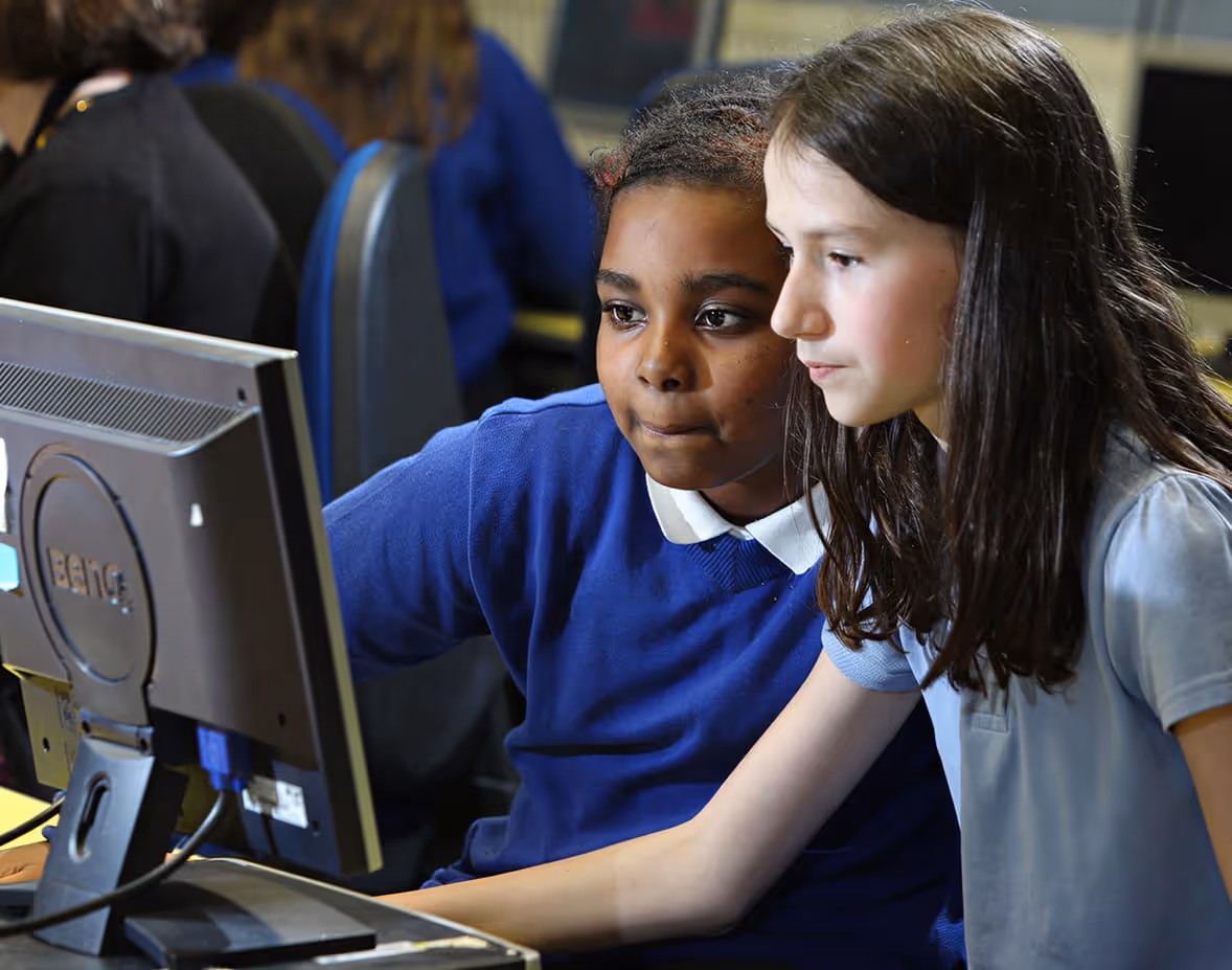 A young girl in a school uniform using a computer mouse with an adult assisting her, while two other children work on computers in the background.