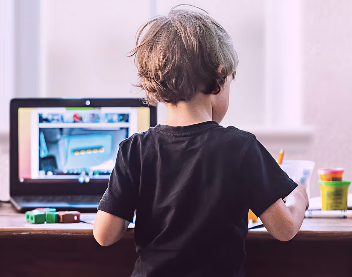 Young boy playing a video game on a computer with a smiling man watching and guiding him in a classroom.