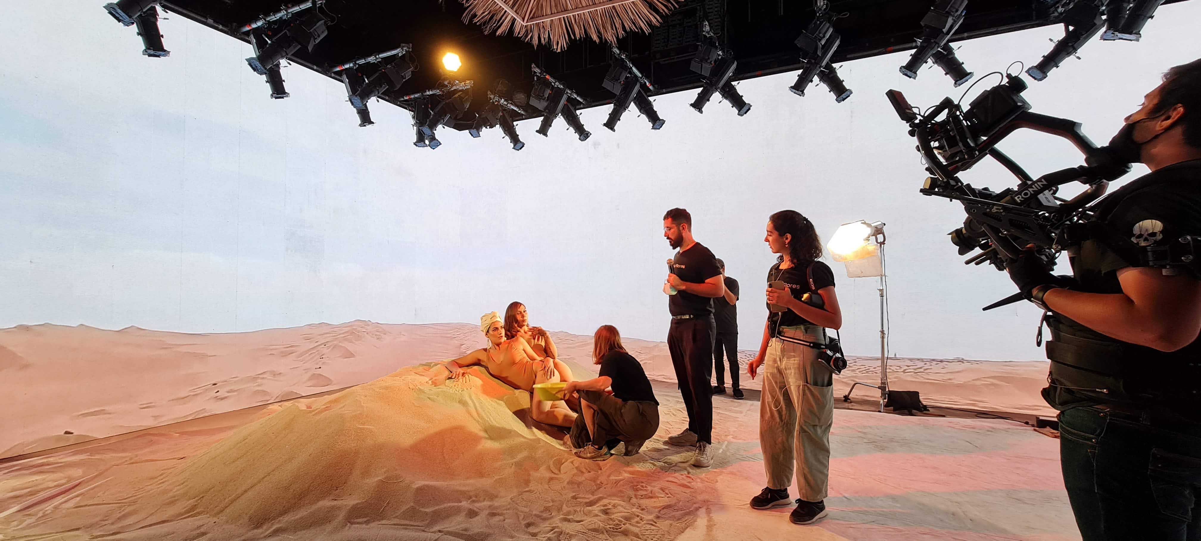 Two women posing on sand with crew and camera filming on indoor studio set with lighting equipment.