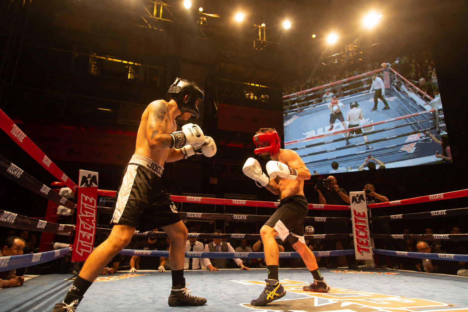 Two boxers wearing headgear and gloves sparring in a ring under bright lights with audience watching.