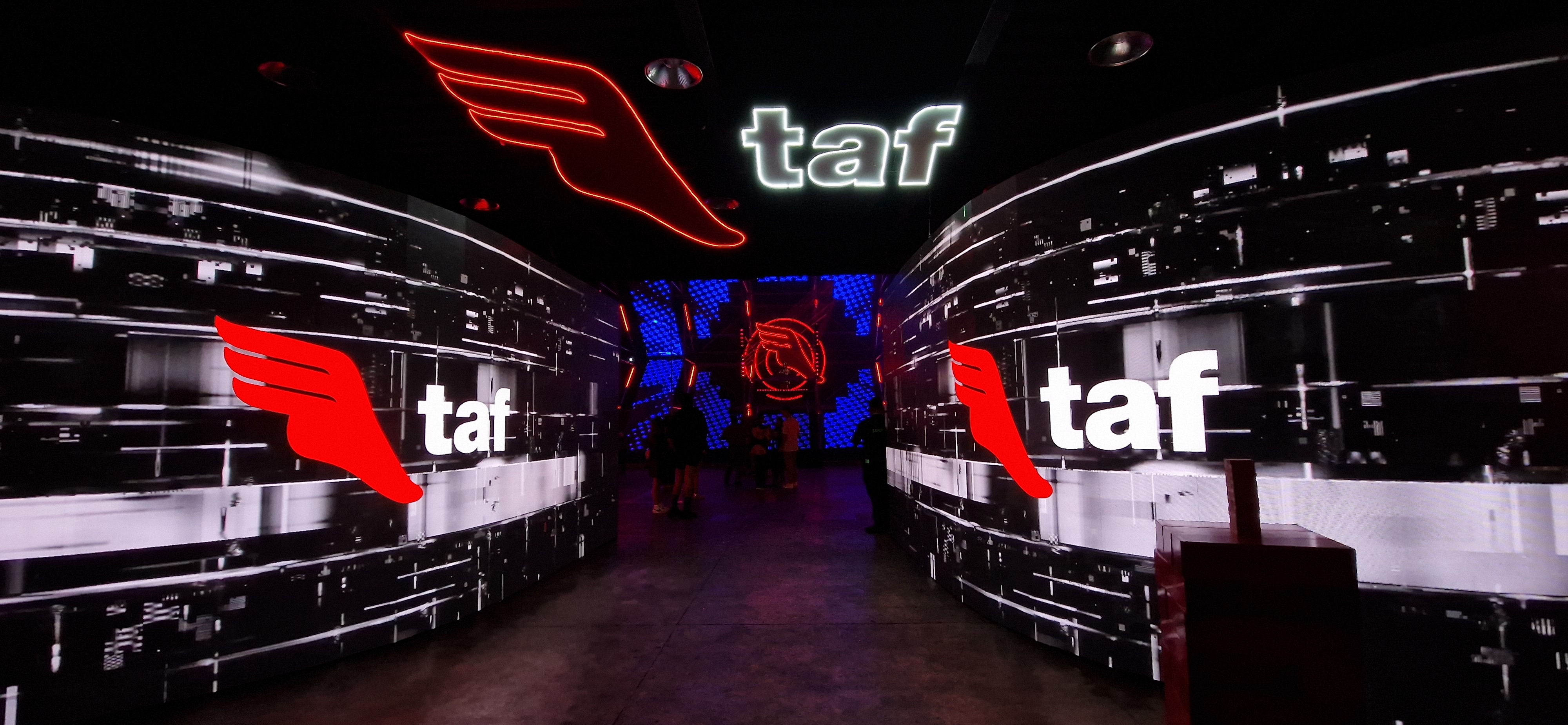 Dark corridor with digital screens showing a red wing and white text 'taf', with people in the distance.