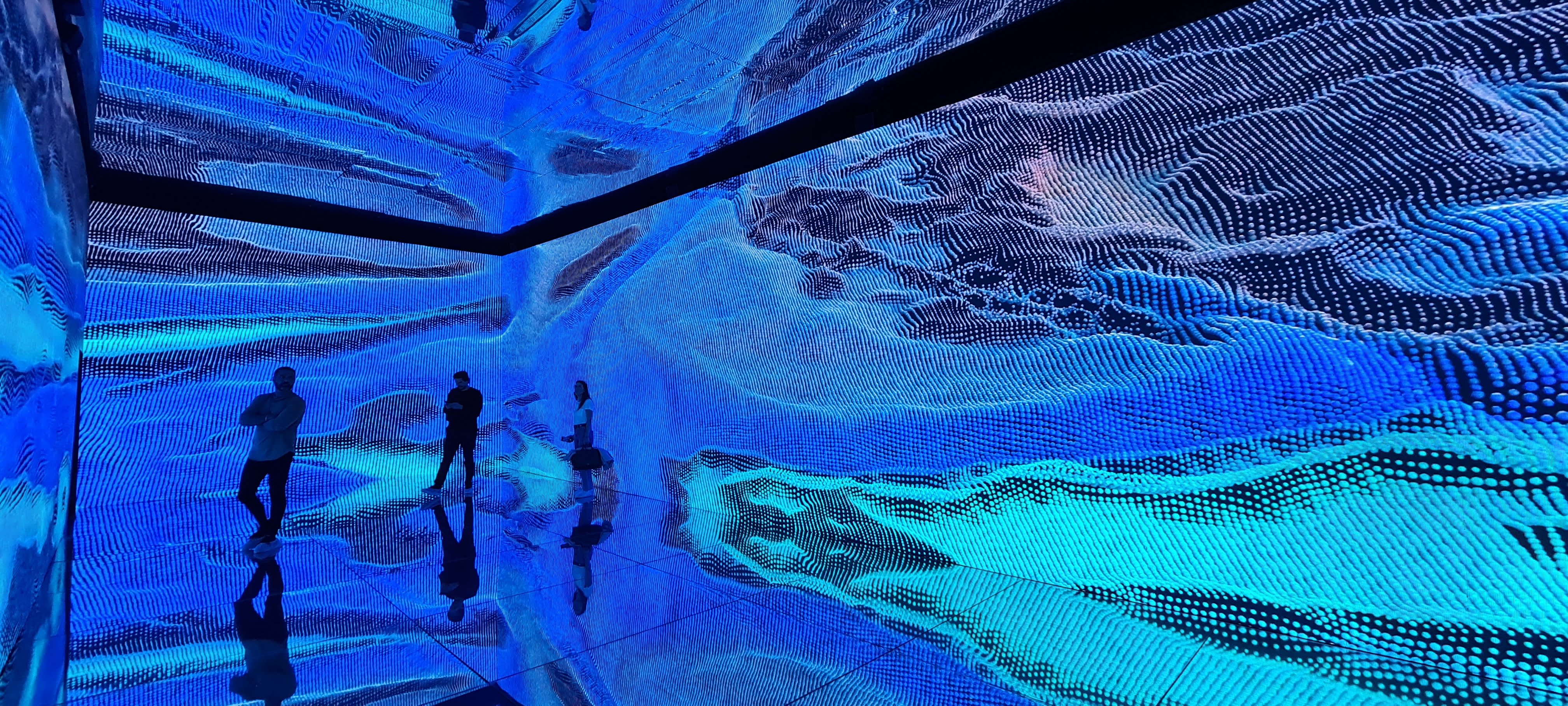 Three people standing in a room with blue digital wave patterns projected on walls and reflective floor.