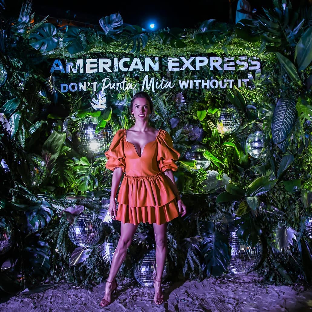 Woman in an orange dress posing in front of green foliage and disco balls with American Express sign.