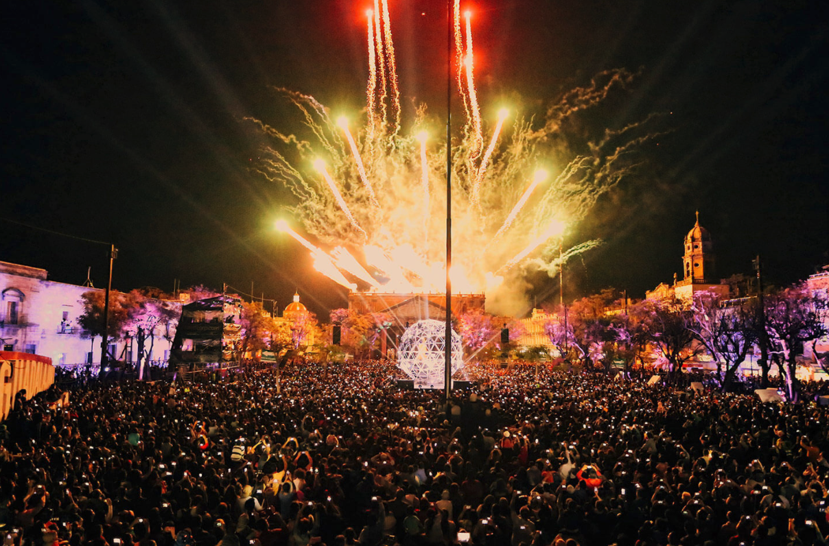 Large crowd watching bright fireworks exploding over a historic city square at night.