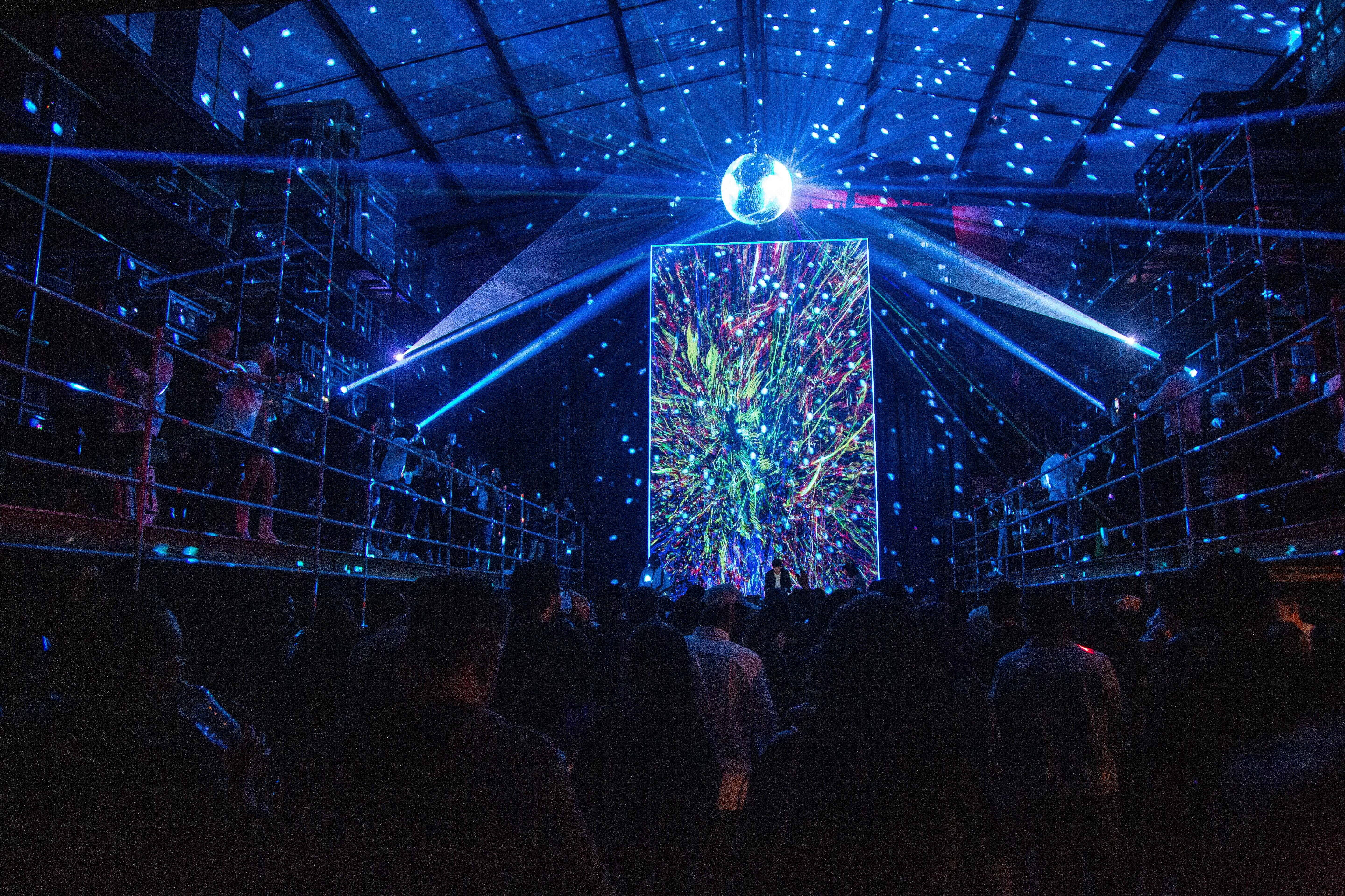 Crowd facing a large colorful digital art screen with disco ball and blue lights in a dark venue.
