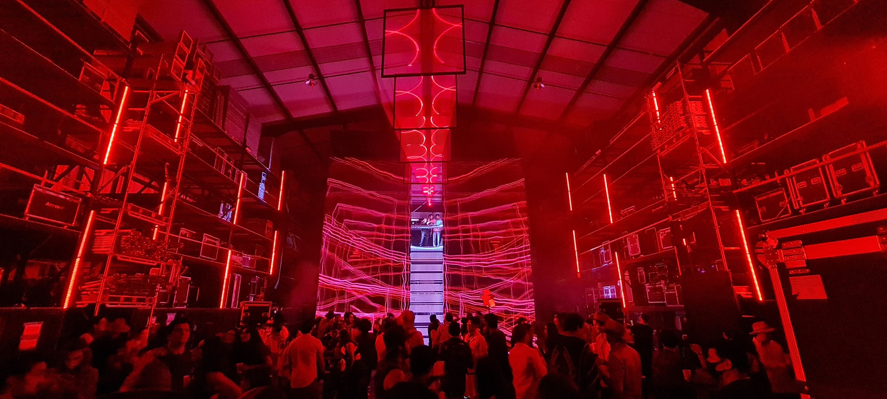 Crowd in a dark venue with red neon lights and a DJ booth elevated in the center background.