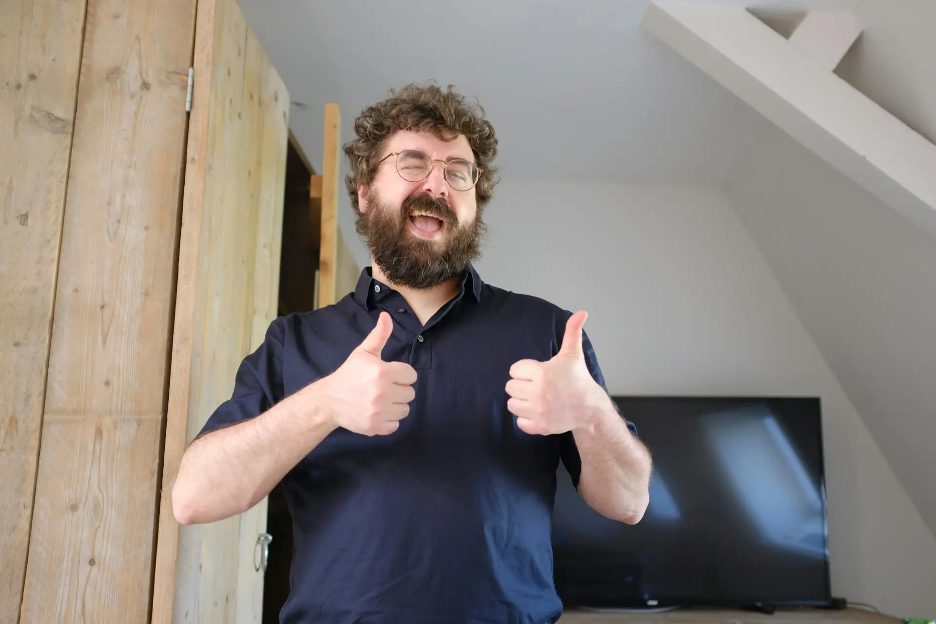 Man with curly hair and beard stands in a room with wooden walls and a TV in the background, smiling and giving 2 thumbs up.