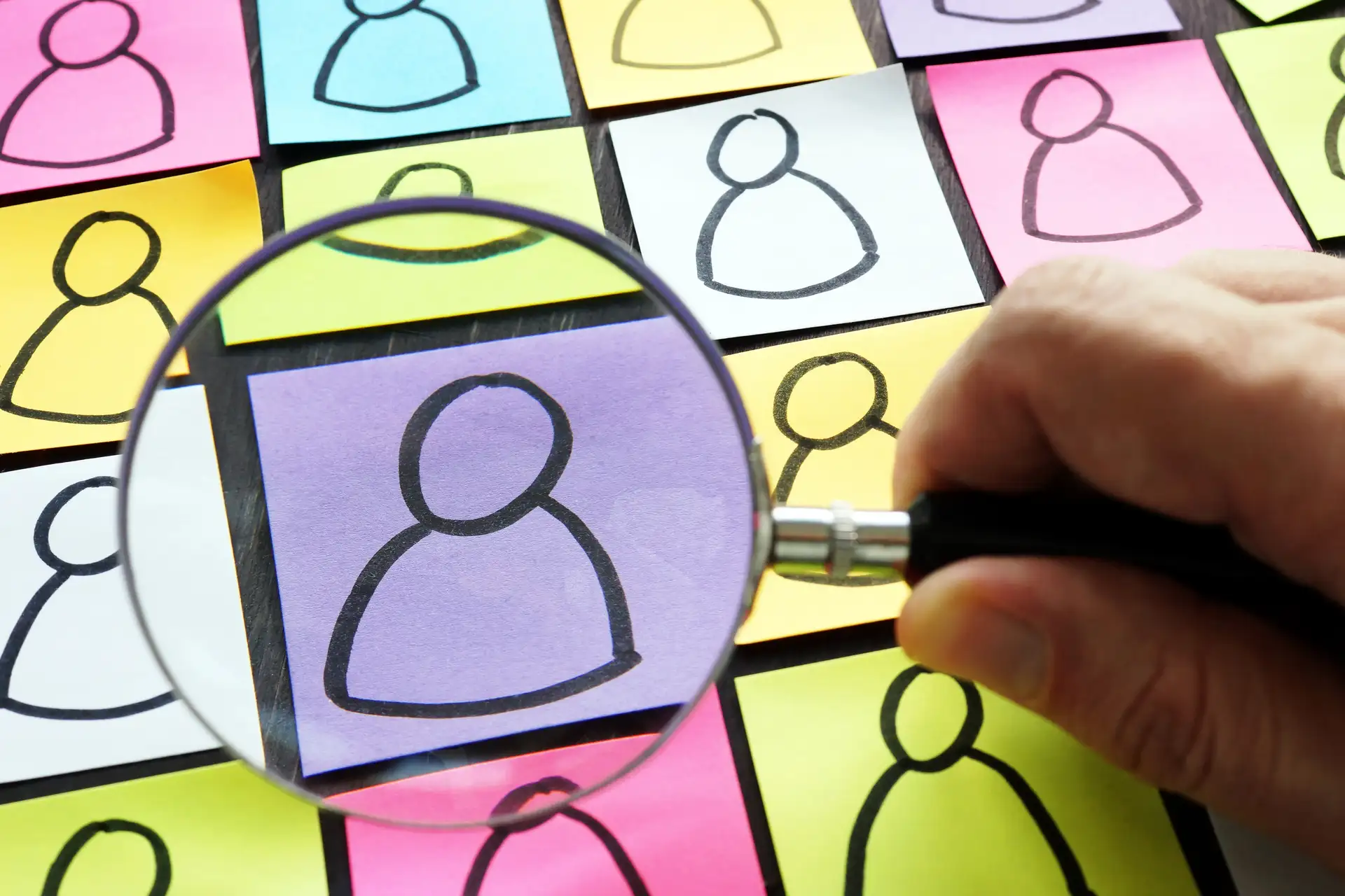 Hand holding magnifying glass over post-it notes with simple stick figure drawings of people on them.