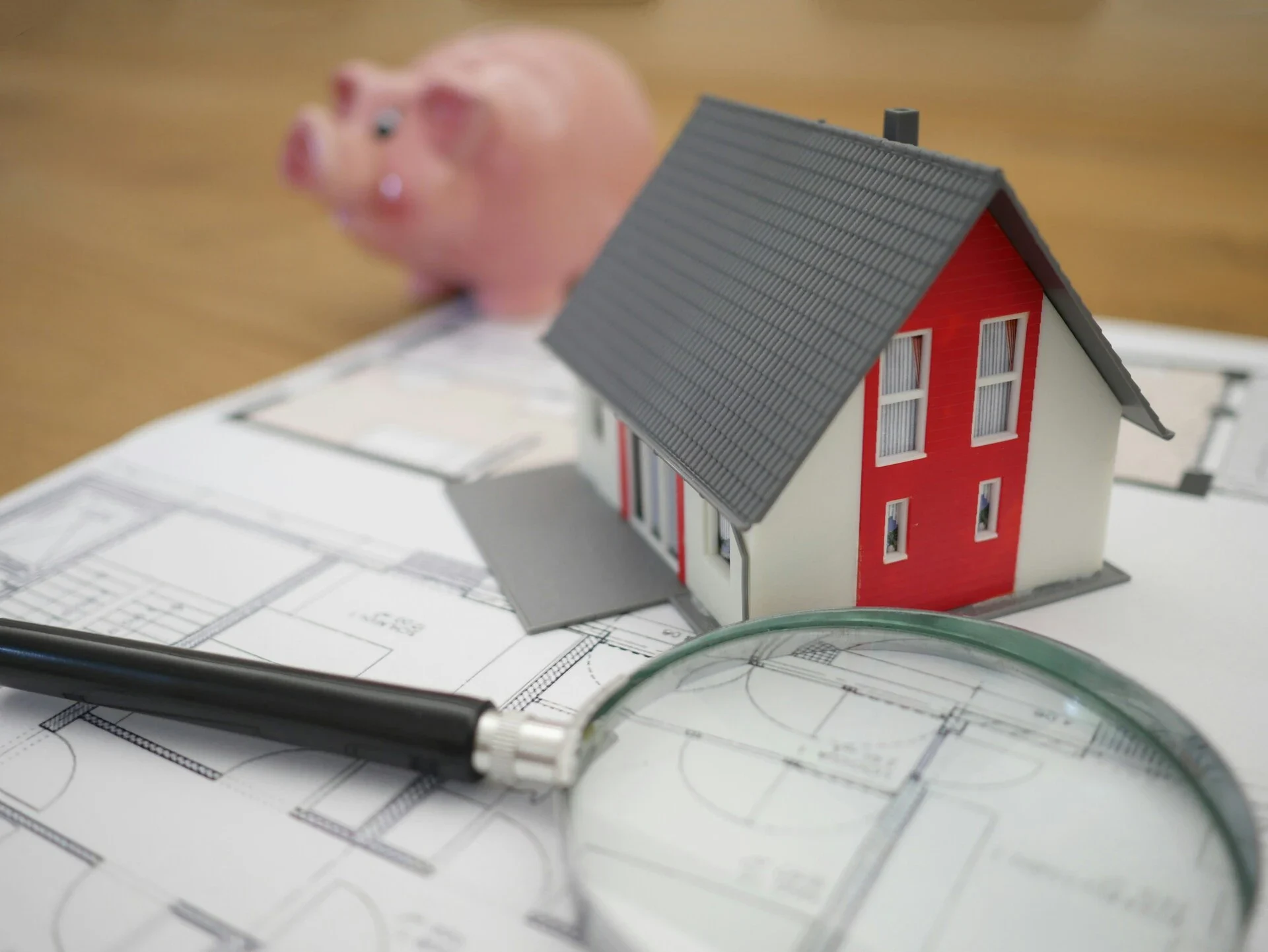 A small house model sits on architectural blueprints alongside a magnifying glass and a piggy bank in the background.
