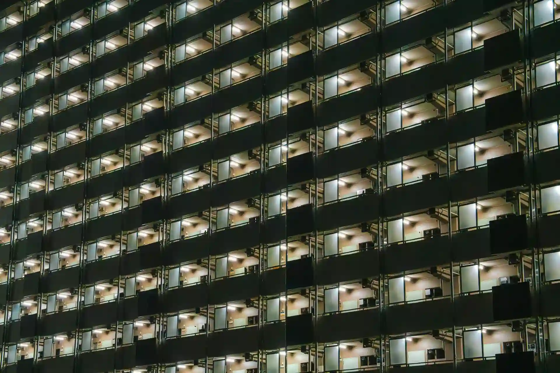 A high-rise building with numerous illuminated balconies in a grid arrangement, viewed at night.