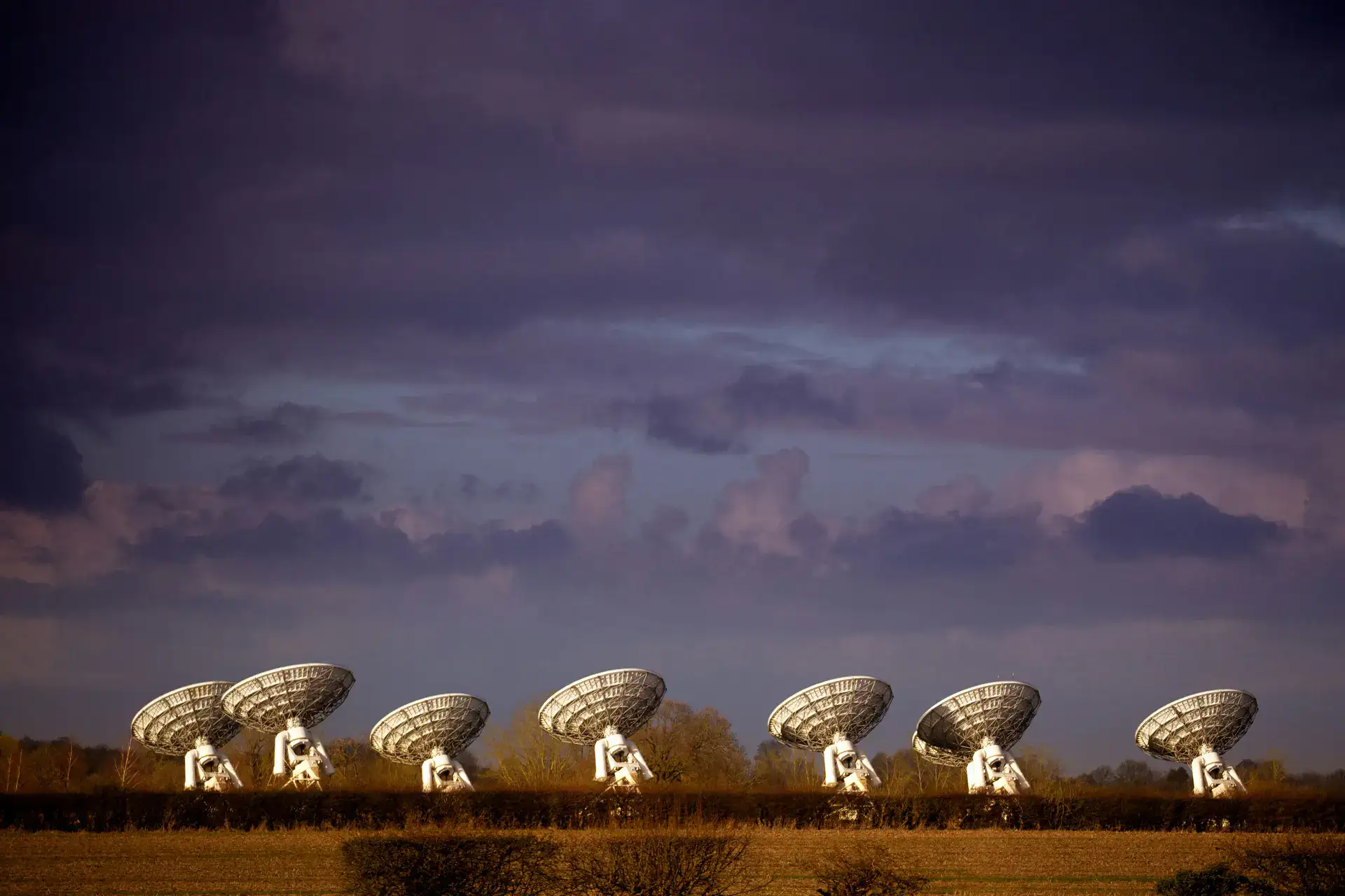 Seven satellite dishes are lined up in a field under a dark, cloud-filled sky at dusk.