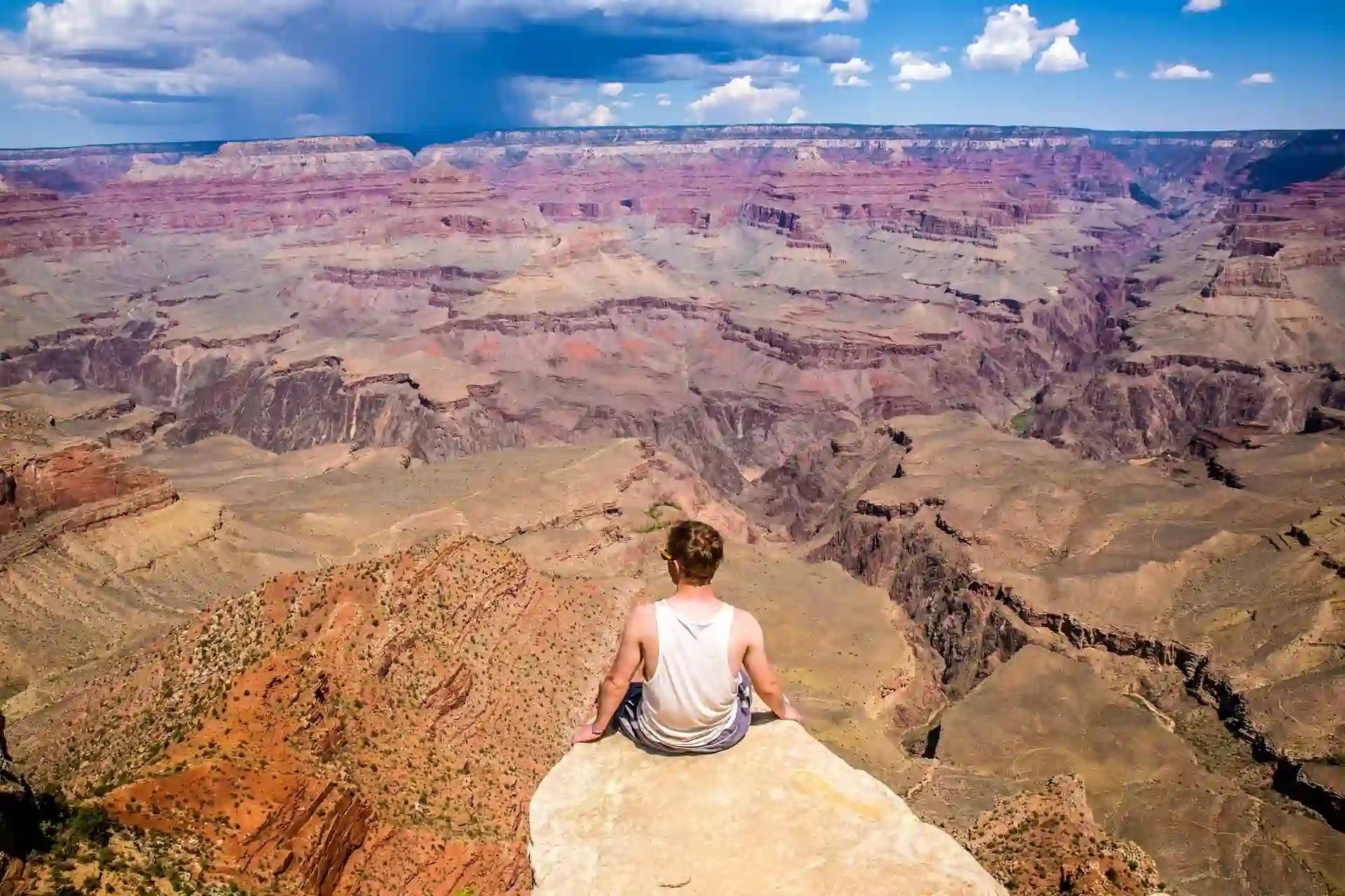 A person sits on a ledge overlooking the vast expanse of the Grand Canyon under a partly cloudy sky.