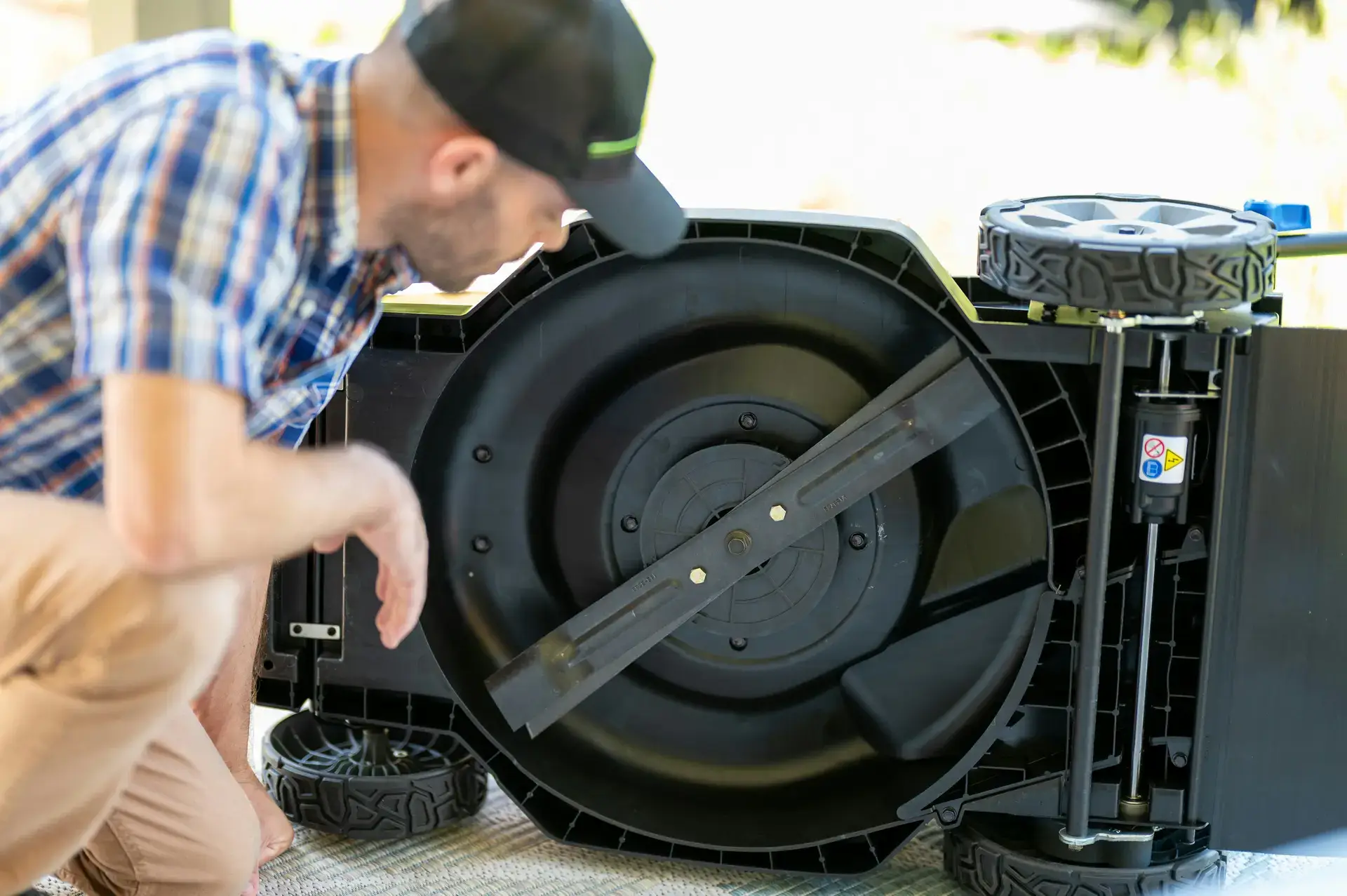 Man wearing a cap inspects the underside of a lawnmower, focusing on the blade.