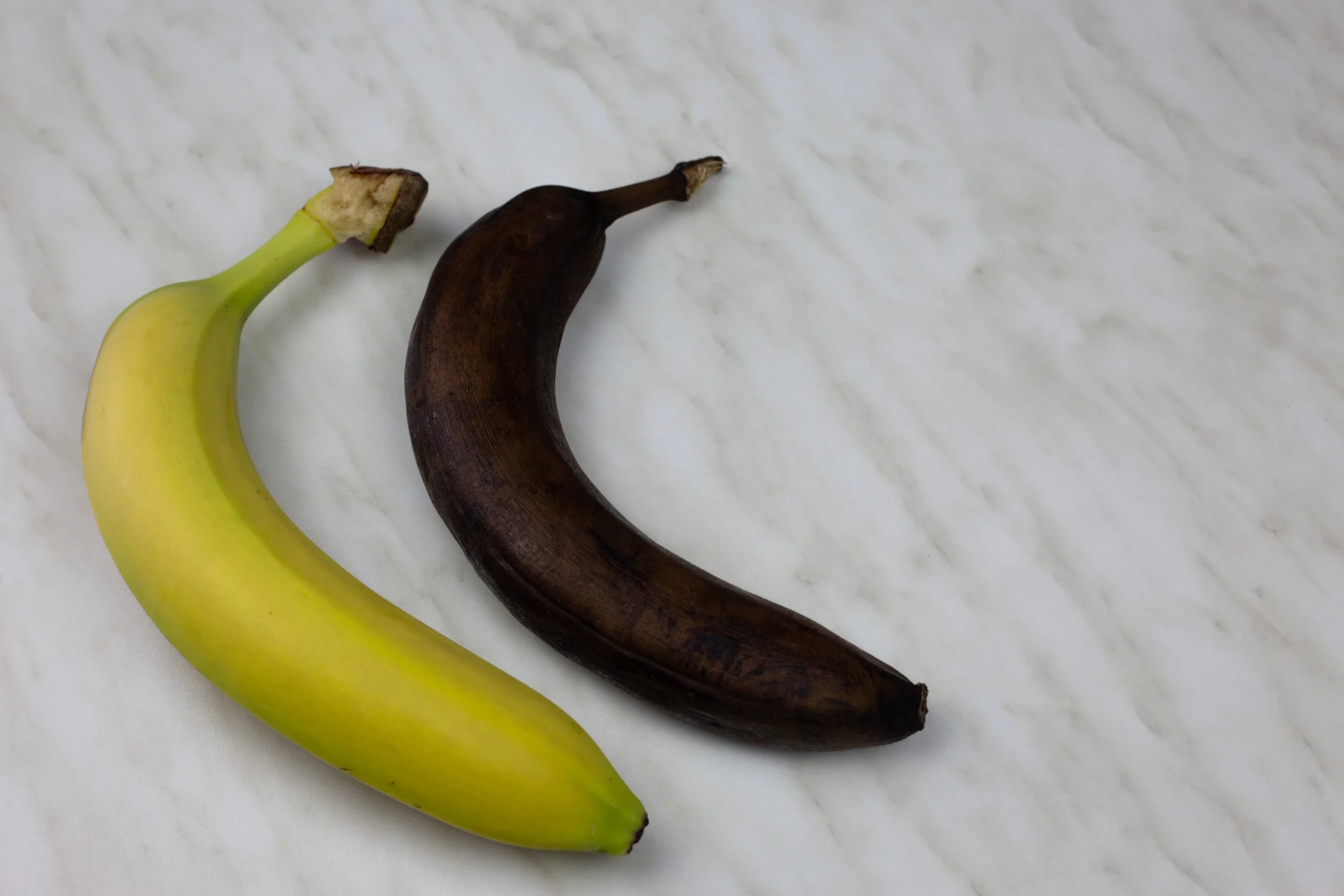 A fresh yellow banana is placed next to an overripe brown banana on a light-colored marble surface for comparison.