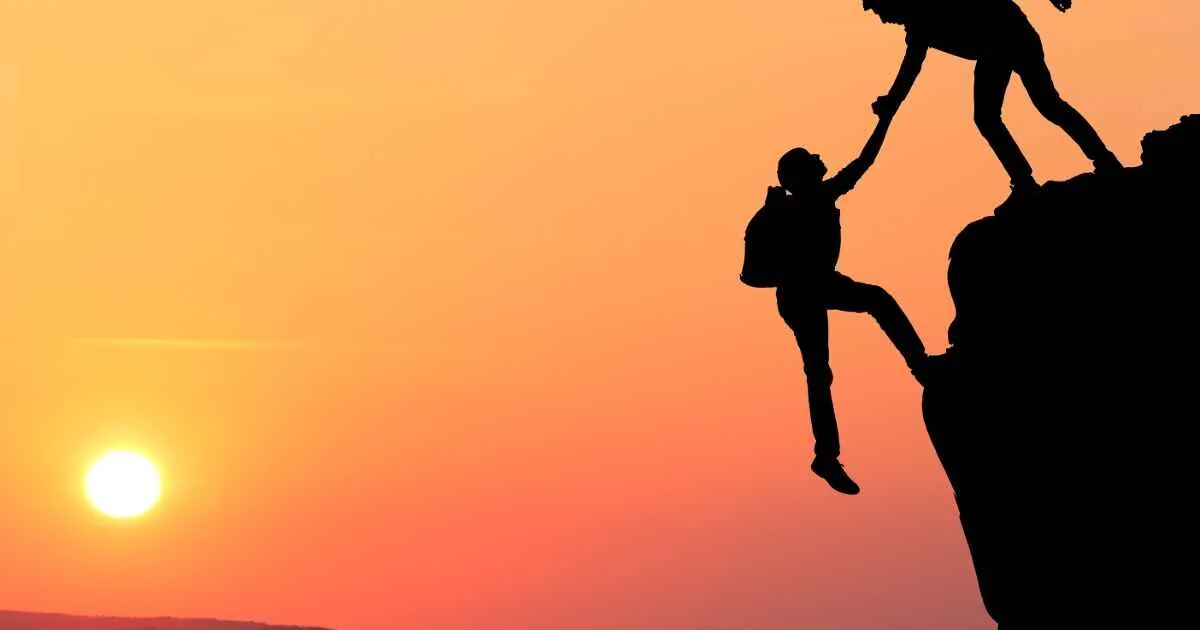 Una persona en silueta ayuda a otra a escalar una roca escarpada al atardecer, encarnando el apoyo y la confianza sin fisuras.