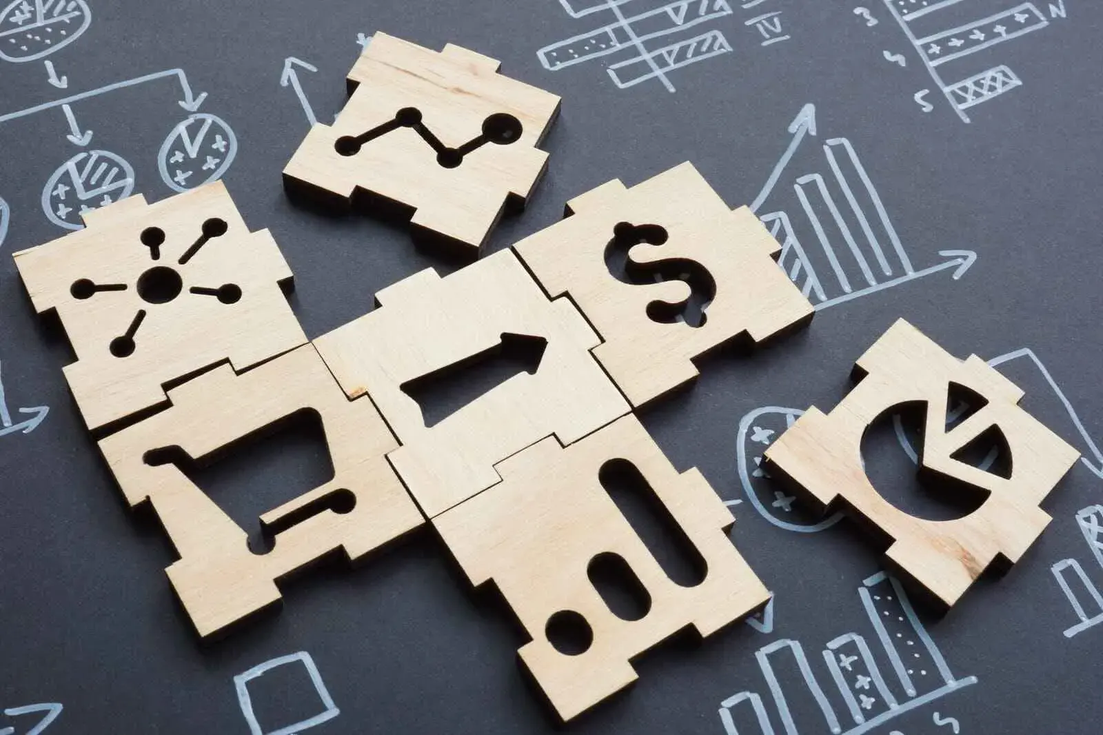 Piezas de rompecabezas de madera con símbolos empresariales como un carrito de la compra, el símbolo del dólar y un gráfico, dispuestos en una pizarra.