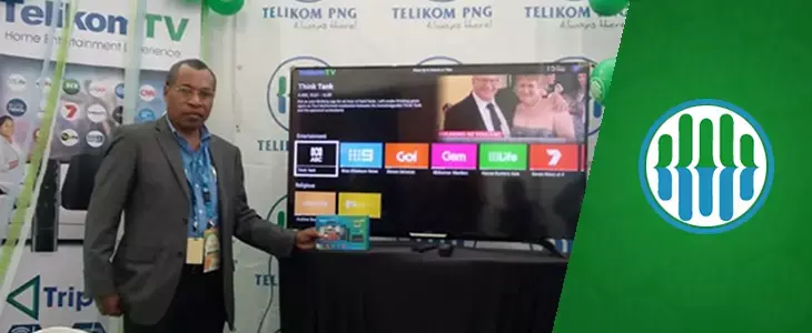Un hombre está de pie frente a una gran pantalla de televisión, sosteniendo una caja con la imagen de un televisor. La pantalla del televisor muestra a un hombre y una mujer.