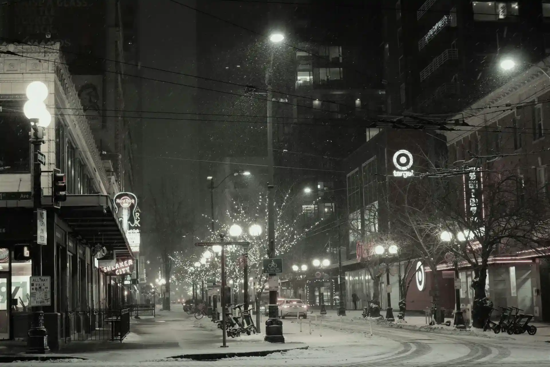Una calle nevada de la ciudad por la noche, bordeada de tiendas y edificios adornados con luces. La nieve cubre el suelo e interrumpe el escaso tráfico.