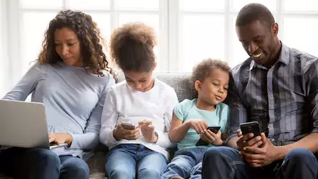 Familia sentada en un sofá. La madre trabaja con el portátil, el padre, la hija y el hijo miran cada uno su teléfono.