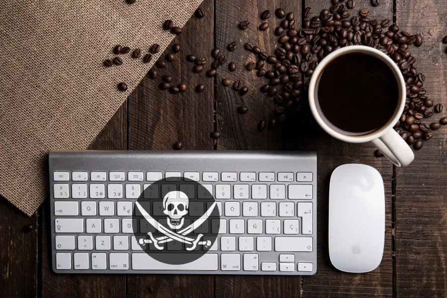 Mesa con taza de café y granos de café alrededor y con ratón de ordenador y un teclado sobre ella. El teclado tiene una bandera pirata en un círculo que representa la piratería en Internet.