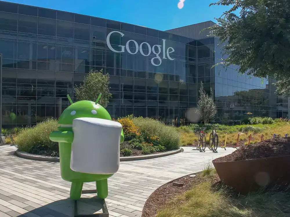 A green google android standing in front of the glass building with google sign on it