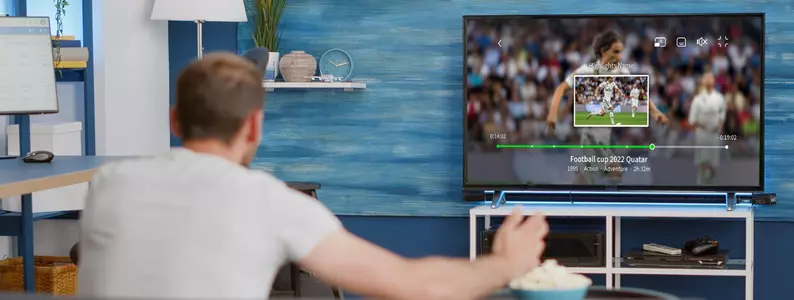 Un hombre está sentado frente a un televisor, viendo un partido deportivo. Sostiene un mando a distancia en la mano, probablemente controlando el televisor. 