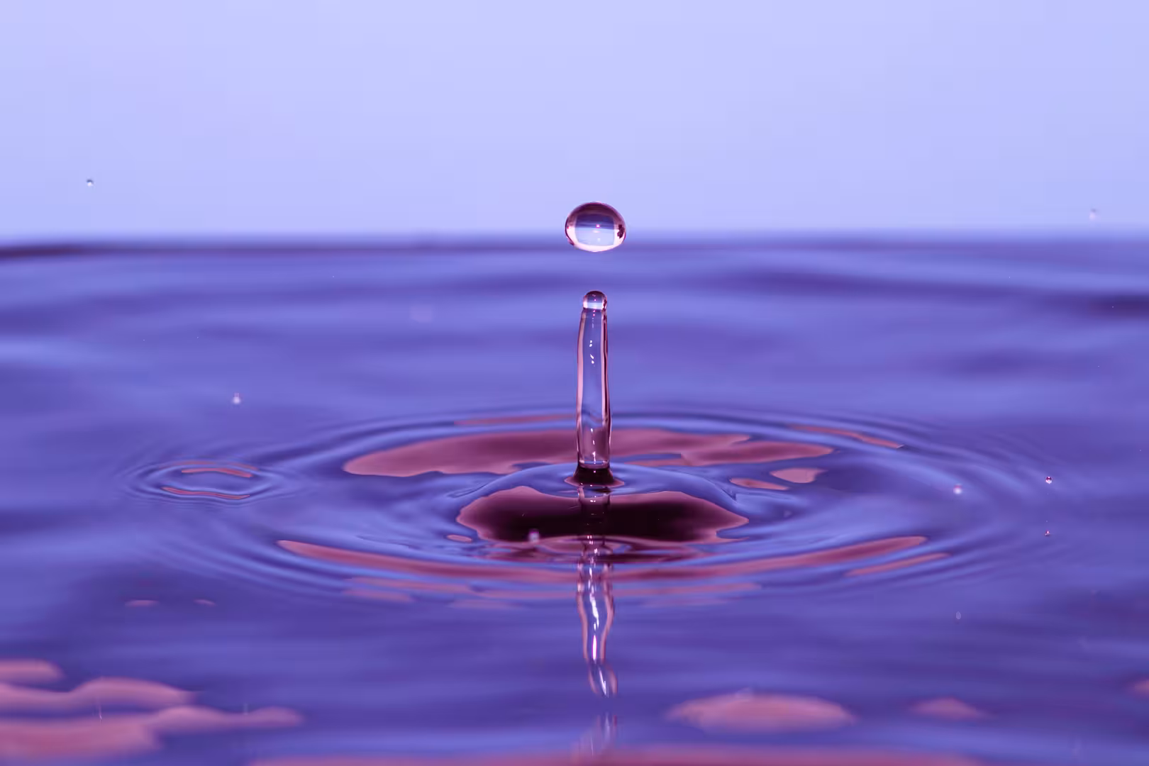 A single drop of water descends into a deep purple liquid, causing a splash and rippling waves.  