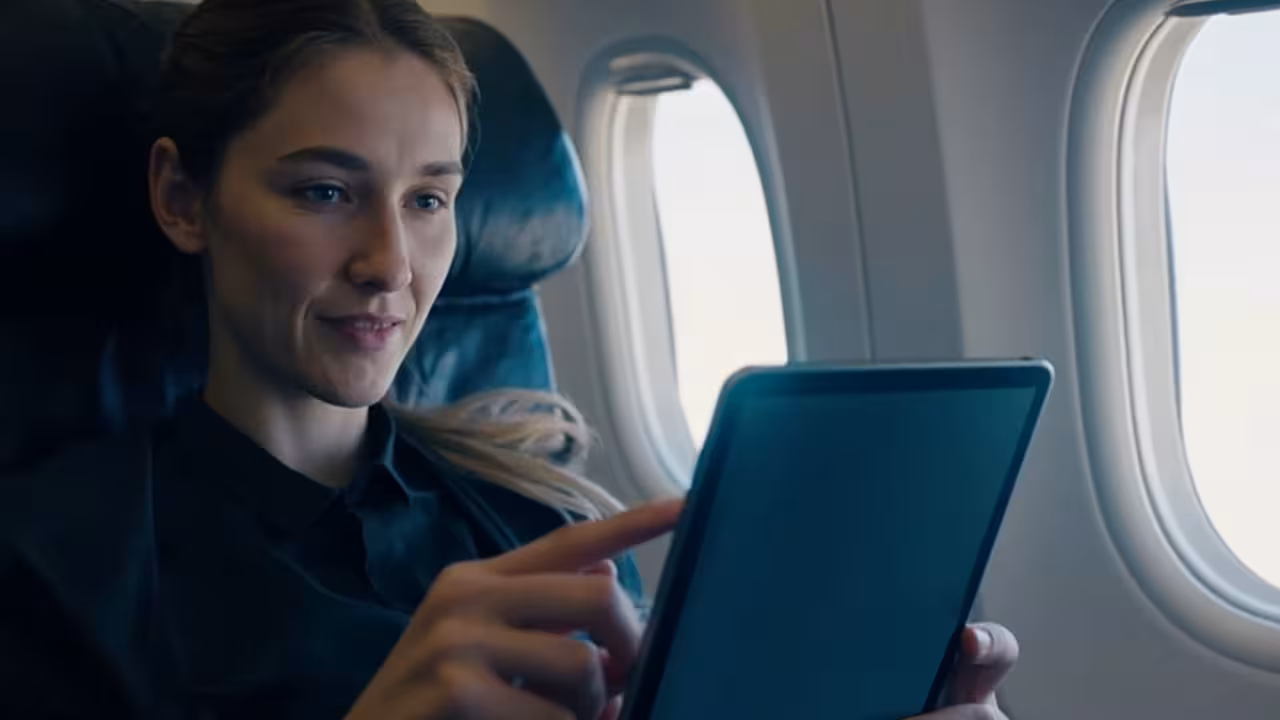 A woman in an airplane is seated comfortably, using a tablet for entertainment during the flight.
