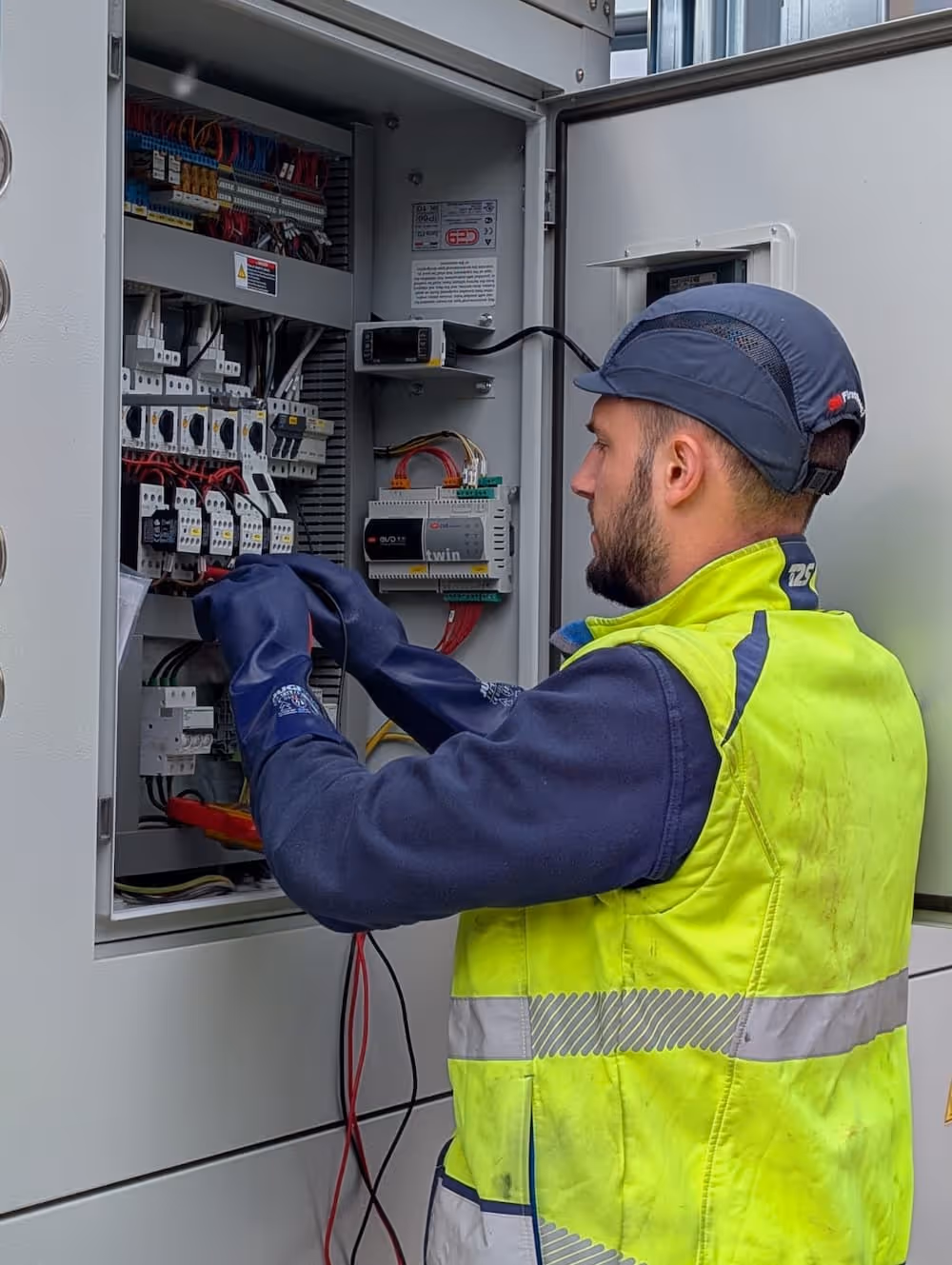 Électricien portant un gilet de sécurité jaune et un casque travaillant sur un panneau électrique ouvert avec câbles et composants.