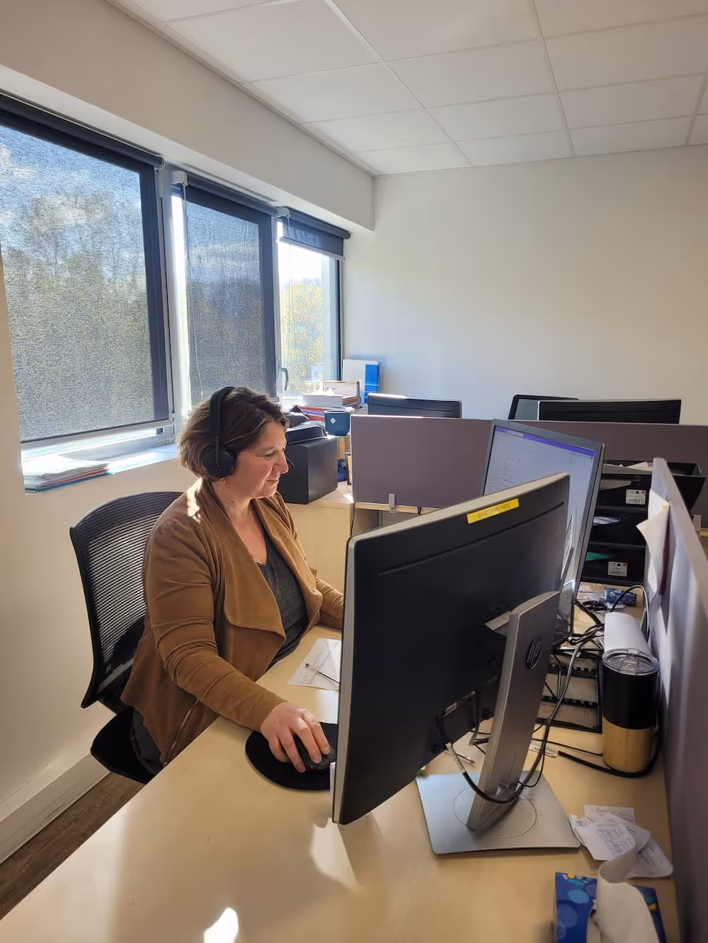 Femme portant un casque, travaillant sur ordinateur dans un bureau lumineux avec grandes fenêtres.