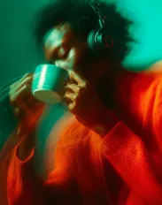 Person with headphones drinking from a cup, wearing an orange sweater, in a greenish ambient light with motion blur effect.