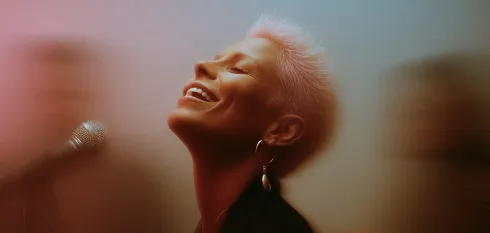 Person with short blonde hair smiling passionately while singing into a microphone with blurred figures in the background.
